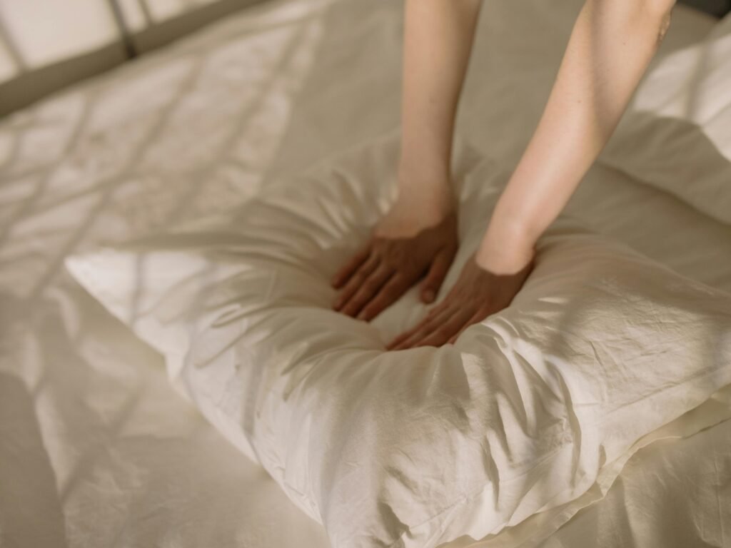 pexels-photo-4108984-4108984 A warm, sunlit bedroom scene with hands fluffing a soft pillow, creating a cozy atmosphere.